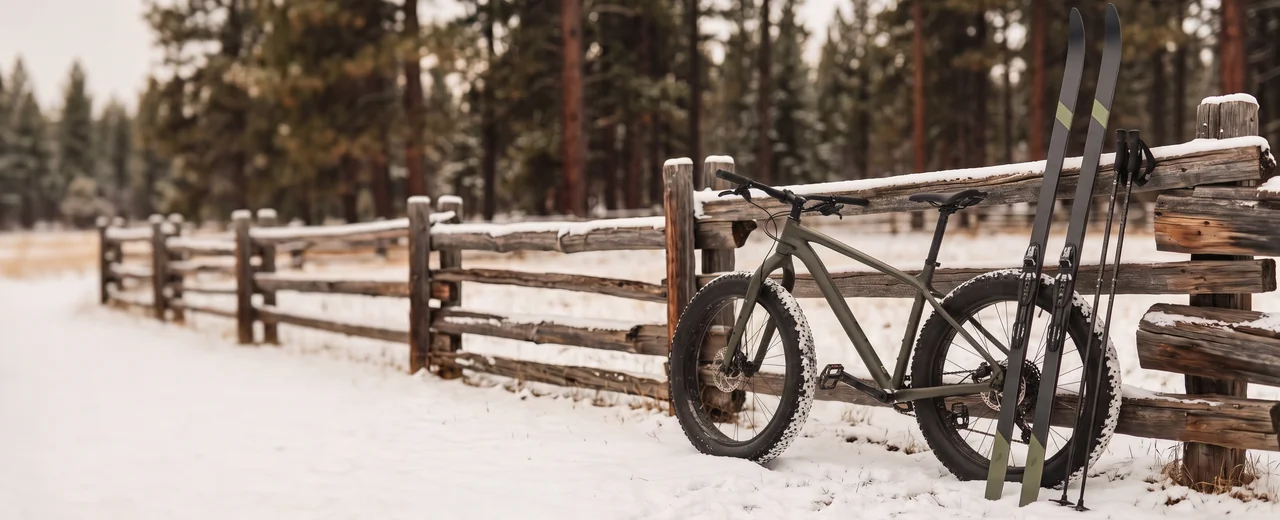 A fat-tire bike and a pair of cross-country skis leaning on a snowy split-rail fence